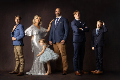 Stylish family portrait captured in a Dallas photography studio featuring a mother in a silver sequin gown, father in a blue blazer, three sons in suits, and a young daughter in a pale blue ruffled dress, all posing against a dark backdrop.