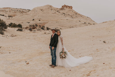 A bride holding her wedding bouquet standing close behind the groom who is wearing a cowboy hat. They are standing in an almost desert-looking landscape located in Capitol Reef National Park.