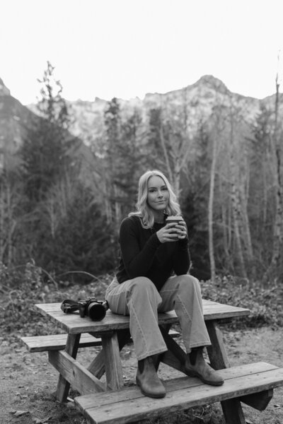 Karli Fisco sitting on a picnic table with her coffee and camera in front of mountains