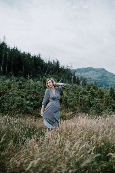 woman is stood in a scenery of mountains and deep dark forests, she is wearing a dress