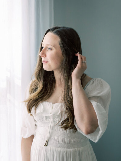 A woman in a white dress standing by a window by Katie Stansfield Photography.