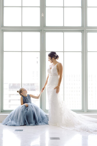 Bride holding hands with a flower girl in a blue dress, standing by a large window, captured by Rodrigo Varela Photography.