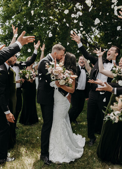 bridal party throwing flowers at couple while they kiss
