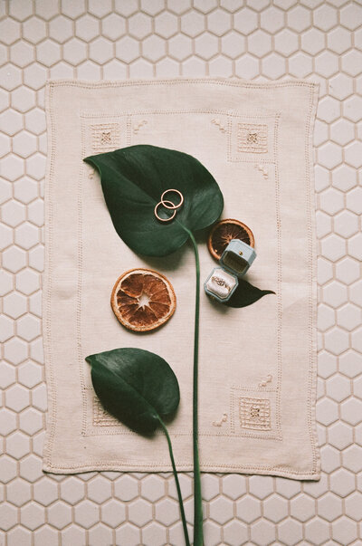 Film photo of a wedding ring flatlay featuring tropical leaves, dried citrus, velvet ring boxes, and a linen mat arranged on hex tile — styled and photographed by a film wedding photographer.