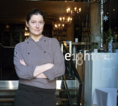 Image: Chef Lia standing with arms folded against a stainless steel counter. Wearing a dark gray Chef’s jacket and black apron.