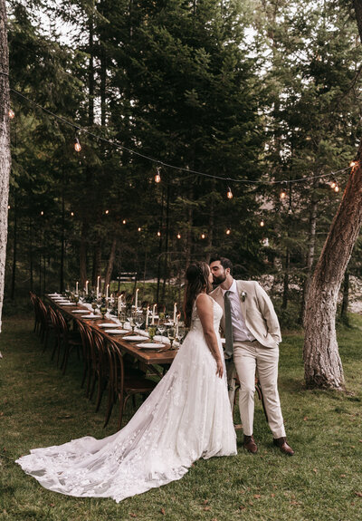 A couple kisses in front of their decorated table in the woods.