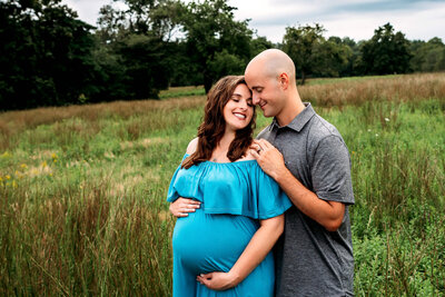 Joyful couple embraces in Connecticut meadow as expecting mother in turquoise off-shoulder gown cradles baby bump during outdoor maternity session with Connecticut maternity photographer