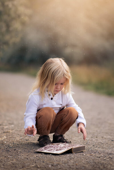 Little boy crouched down opening an old book with dust falling on him.  Portrait by Brittany Danielle Photography