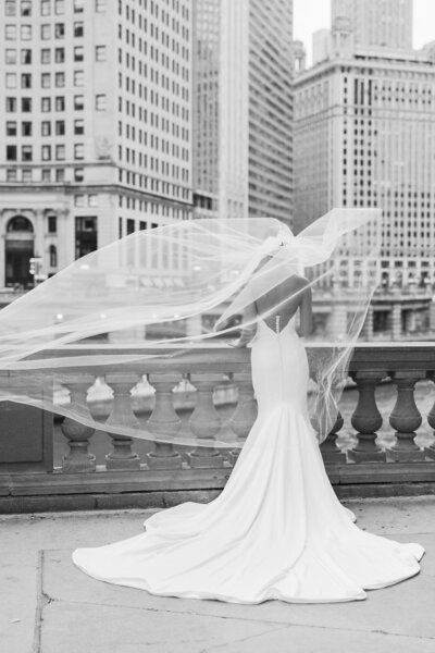 A timeless black and white wedding portrait of a bride facing away as her veil lifts in the wind, capturing a soft and candid moment.