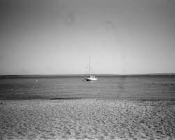 Black and white photo of a tranquil beach scene. A lone sailboat floats on calm water under a clear sky, creating a serene, peaceful atmosphere.