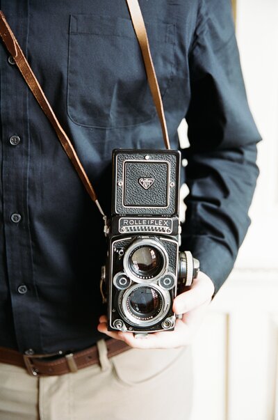 Devon wearing a black button up holding his Rollieflex 2.8F Twin Lens Reflex film camera in his left hand