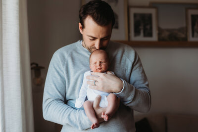 Dad holding newborn baby against his chest in living room for at home newborn session