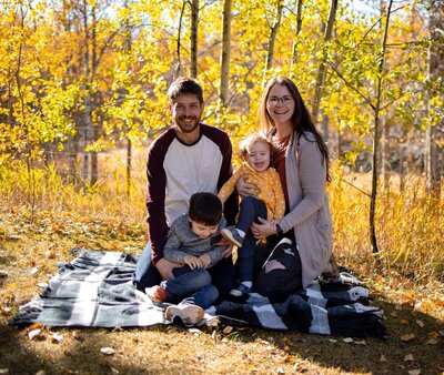 A family of four sit on a picnic blanket in front of yellow leaves. The dad is on the back left, the mom is on the back right, and their son and daughter sit in front of them