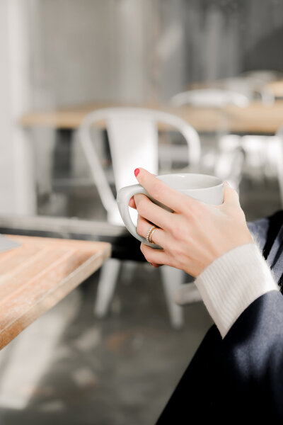 Close-up of a woman's hands holding a coffee mug, with sunlight streaming into a modern café, symbolizing a moment of focus and clarity for entrepreneurs seeking simplified systems and streamlined workflows