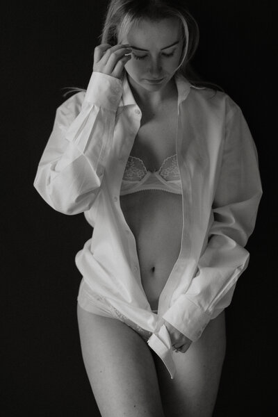 Black and white photo of woman in lingerie and a button up shirt standing in front of a black wall