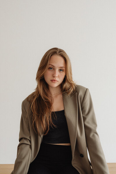  A young woman with light auburn hair wears a dark beige blazer over a smart black cropped bandeau top and high-waisted black trousers. She looks directly at the camera with a poised expression, leaning on a wooden table.