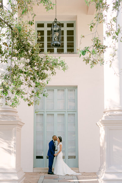 Bride and groom share a kiss in an elegant outdoor setting in Miami, Florida, surrounded by greenery and classic architecture, captured by Rodrigo Varela Photography. Romantic wedding portrait showcasing timeless love and refined style.