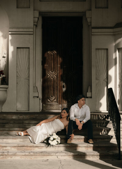 A young couple sits on sunlit stone steps outside a building with ornate wooden doors. The woman in a white dress reclines elegantly, holding flowers.