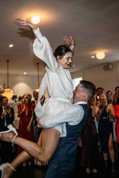 Bride in a white dress being lifted joyfully during a high-energy wedding reception dance, surrounded by cheering guests