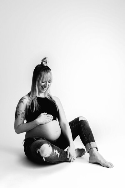 black and white image of a pregnant woman wearing jeans and a bralette sitting on the floor