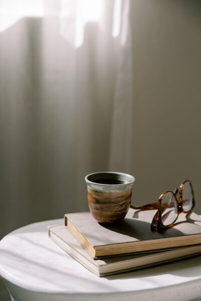 A coffee mug and pair of glasses sitting on 2 books on a table 