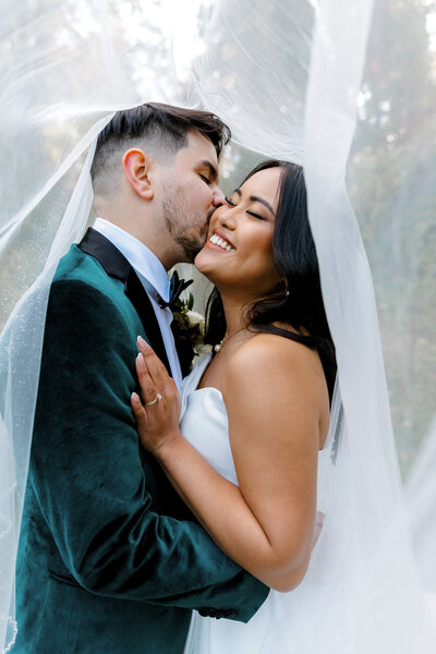 bride and groom under veil getting kissed on the cheek