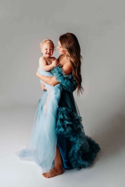 A low key motherhood portrait captured by Mobile, AL family photographer of a mom wearing a blue tulle gown while holding her giggling toddler who is wrapped in blue tulle fabric. The mother looks at her child happily, as the child makes eye contact with the camera.