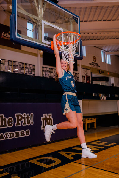 A senior basketball player hangs from the basketball hoop rim as she smiles at the camera.