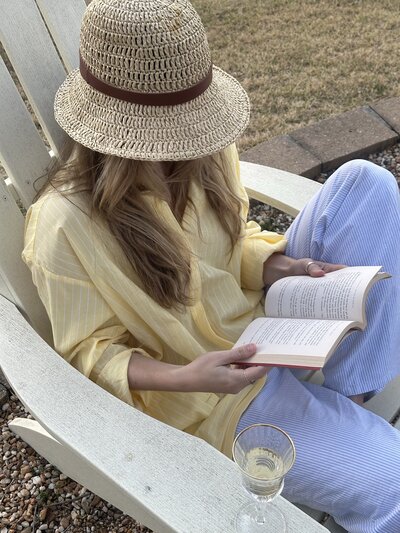 A woman in a straw hat and yellow shirt sits in a chair outdoors, reading a book. Her relaxed pose and the wine glass convey a tranquil mood.