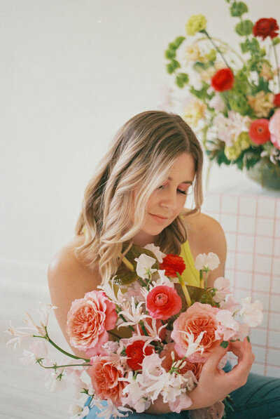 Portrait of a florist. A blonde haired white woman holding a bouquet of pink and white flowers. She is looking down at the bouquet and her hair is slightly falling in her face. The background is mostly white with another flower arrangement behind out of focus.