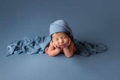 Newborn photography — baby boy in soft blue wrap and matching bonnet posed on blue backdrop with hands under chin