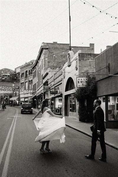 In Bisbee, Arizona, a marrier twirls in their wedding dress in the middle of a historic street while their partner looks on, capturing a joyful moment.