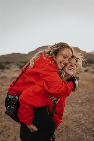 Two individuals wearing matching red jackets share a joyful moment in a desert landscape, one giving the other a piggyback ride as they both laugh with pure happiness.