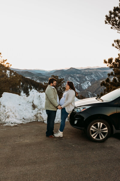 Expecting Couple hold hands and lean on a red car with the Rocky Mountains behind them