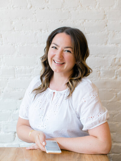 A woman with wavy brown hair smiles while holding a smartphone in her hand. She is wearing a white blouse with delicate embroidery, sitting at a wooden table, and the background features a light brick wall.