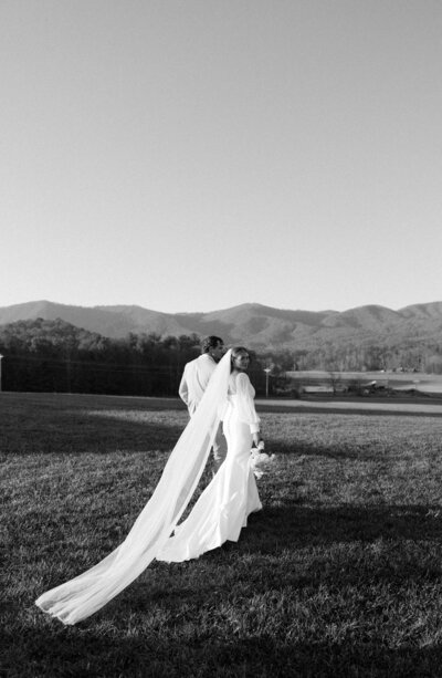 A bride smiles over her shoulder while walking hand in hand with her groom in an open pasture at sunset