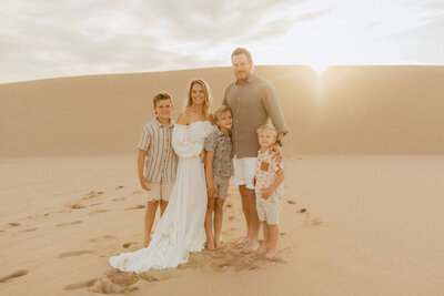 A family with 3 young boys stands on the beach  and pose for a family photo. Everyone is in nice clothes in soft neutral colors