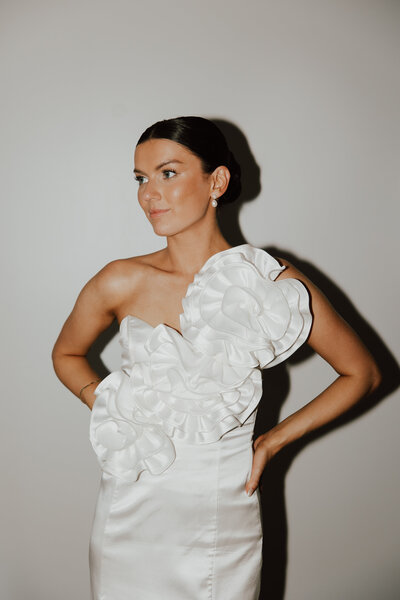 Bride in an asymmetrical white gown with dramatic floral detailing, wearing a sleek low bun styled by Promise Artistry for a timeless, modern bridal look.