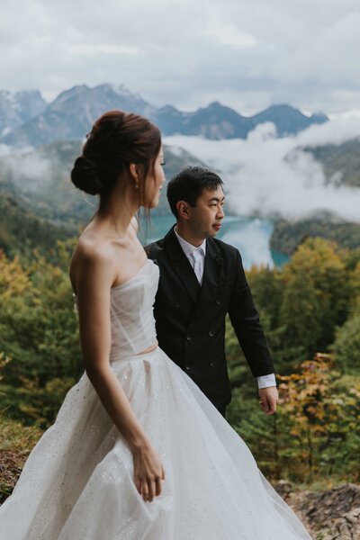 A lesbian couple holds hands facing each other, kissing under a mountain peak in Slovenia. They are both wearing white wedding attire.