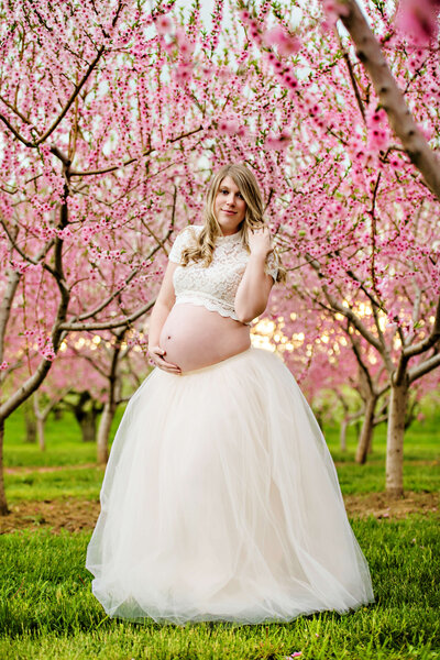 Blonde pregnant lady standing in the cherry blossoms at sunset wearing a cream tutu and lace crop shirt for her outdoor maternity photography session.