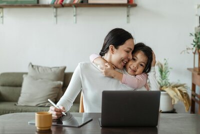Mom at her desk with laptop while daughter hugs her. 