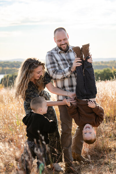 a smiling dad holds his young son upside down as the mom tickles him. the family all laughs together as they stand in a softly lit grassy field
