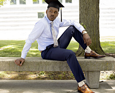 Senior boy in light blue dress shirt, tie, navy blue dress pants, and brown dress shoes sitting on concrete bench outside of the DIA in Detroit, Michigan
