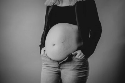 Een zwangerschapsshoot in de studio door Evita Fotografie. Een vrouw met de handen in haar zak met een zwart jasje en hemdje aan met een blote buik