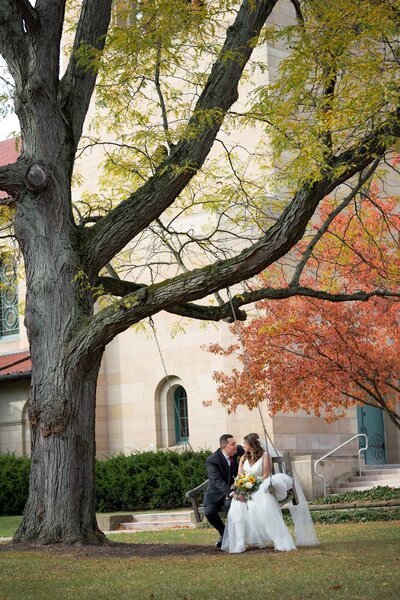 Bride and groom swing together underneath a tree with fall folliage