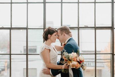 Newlyweds touch foreheads while standing in an industrial window
