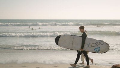 The couple is waking on the beach with the surfing board on hands of a man while holding their hands together. filmed by Jimmy Shin Films