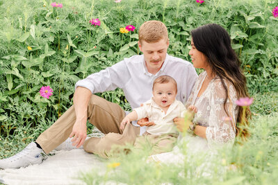 A young mother and father sit on a blanket with their baby son. Photo taken at a wildflower field near the Outer Banks.