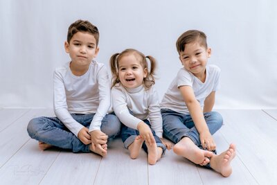  A group portrait of three siblings sitting side-by-side on a light-colored wooden floor against a white background. The older brother on the left has a neutral expression, while the younger brother on the right is smiling. The little girl in the middle, with her hair in two pigtails, is laughing with her mouth open. All three are wearing white long-sleeved shirts and blue jeans.