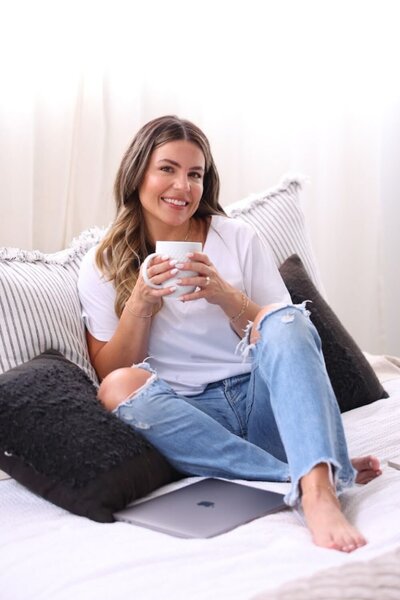 Natalie holding a white mug and smiling warmly while sitting on a cozy bed with her laptop—inviting calm, clarity, and connection.